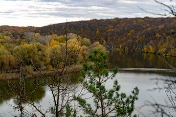 Autumn trees with yellow and red leaves on a hill above calm river reflecting forest and cloudy sky. Rural landscape photography. Autumn season and nature concept.