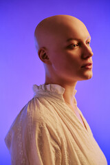 Caucasian young adult woman with alopecia standing in profile against gradient background, showing smooth bald scalp and delicate facial features, wearing light textured blouse
