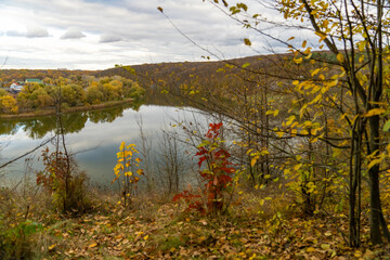 Autumn trees with yellow and red leaves on a hill above calm river reflecting forest and cloudy sky. Rural landscape photography. Autumn season and nature concept.