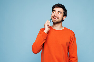 Young fun man he wear orange sweatshirt casual clothes talk speak on mobile cell phone conducting pleasant conversation isolated on plain pastel light blue cyan background studio. Lifestyle concept