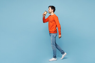 Full length young man he wear orange sweatshirt casual clothes hold takeaway delivery craft paper brown cup coffee to go isolated on plain pastel light blue cyan background studio. Lifestyle concept.