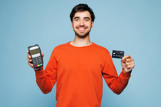 Young man he wearing orange sweatshirt casual clothes hold wireless modern bank payment terminal to process acquire credit card isolated on plain pastel light blue cyan background. Lifestyle concept.