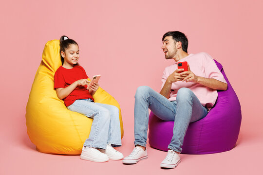 Full body young parent man with child girl wear red casual clothes Daddy little kid daughter sit in bag chair use mobile cell phone isolated on plain pink background Father's Day Love family concept - Powered by Adobe
