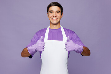 Young smiling happy satisfied man wear violet t-shirt apron while doing housework tidy up showing thumb up like gesture isolated on plain pastel light purple background studio. Housekeeping concept.