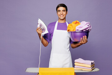 Young smiling man he wear violet t-shirt iron clothes on ironing board while doing housework tidy up look camera isolated on plain pastel light purple background studio portrait. Housekeeping concept.