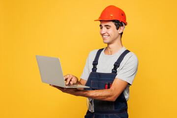 Young employee laborer handyman IT man wear helmet overall use work on laptop pc computer isolated on plain yellow background. Instruments accessories renovation apartment room. Repair home concept.