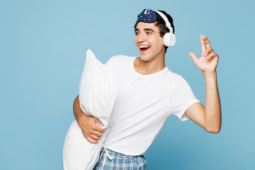 Calm young happy man he wear pyjamas jam sleep eye mask rest relax at home hold pillow listen to music on headphones isolated on plain pastel light blue cyan background. Good mood night nap concept.