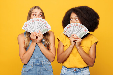 Two young friends buddies women they wear denim casual clothes together hold in hand fan of cash money in dollar banknotes isolated on plain yellow orange background studio portrait Lifestyle concept