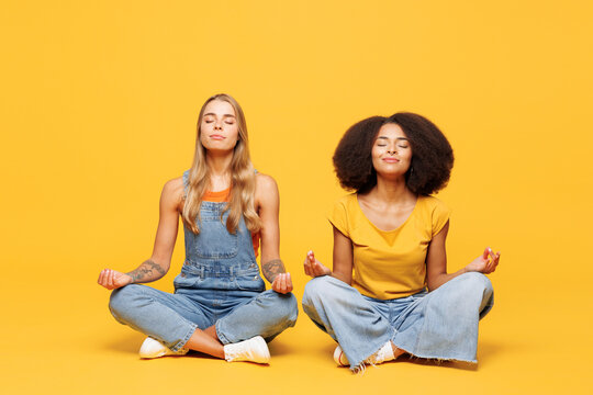 Full body two young friends buddies women wear denim casual clothes sit together hold spreading hands in yoga om aum gesture relax meditate try to calm down isolated on plain yellow orange background.