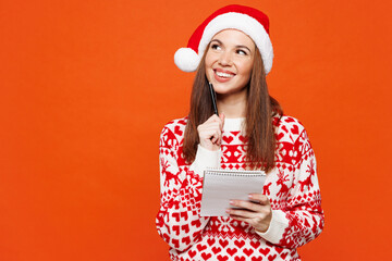 Merry minded pensive young woman wear red warm knitted Christmas sweater Santa hat posing hold notebook making wish list isolated on plain orange background Happy New Year celebration holiday concept