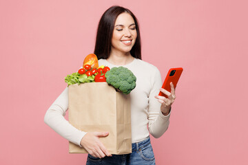 Young smiling happy woman she wear white casual clothes hold brown craft bag for takeaway mock up with food products use mobile cell phone isolated on plain pink background Delivery service from shop