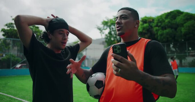 Hispanic man showing phone to friend on soccer field, explaining recent moves or highlights while other reacts in disbelief and surprise, expressing shock and excitement outdoors