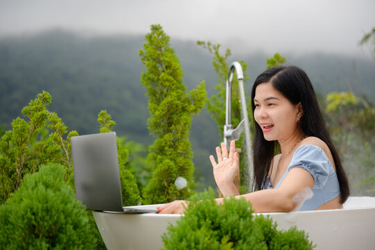 A happy lifestyle in Thailand, the idea of remote communication, and a smiling Asian woman waving her hand while video calling on a laptop in an outdoor bathtub with a foggy mountain view.