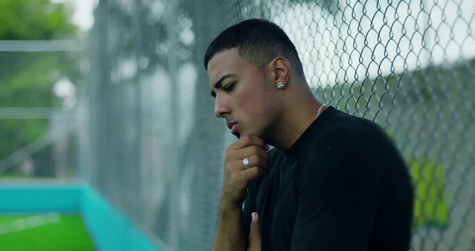 Latin Hispanic man of African descent leaning against chain-link fence with hand on chin, appearing thoughtful and pensive, reflecting quietly in moment of introspection