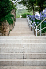 Obraz premium Long outdoor staircase made of light-colored stone leading upward and bordered by white metal railings. There are green trees and a hint of distant mountains on the background. Atmosphere is calm