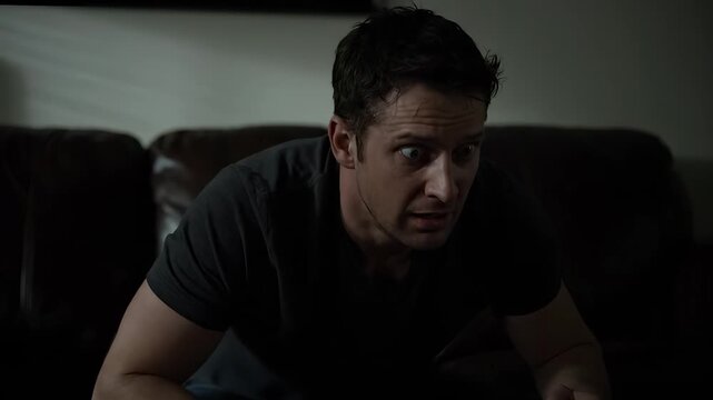 man sitting on couch in livingroom watching television. shirt and jeans worn. hand clenched into fist on knee. darkness surrounds area. floor lamp off. cushion indented by weight. silhouette form.