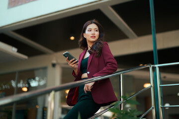 Asian businesswoman browsing smartphone looking away from railing