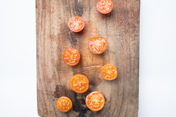 Top view flat lay of fresh sliced cherry tomatoes scattered on a rustic wooden cutting board. The board is on a clean white background, providing copy space.
