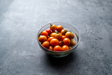 A clear glass bowl filled with fresh, ripe cherry tomatoes. The bowl rests on a dark, textured slate background. Minimalist food concept with copy space.
