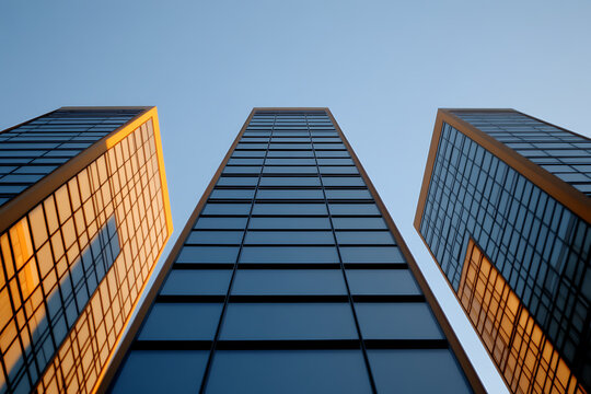 Looking up at modern office towers against a clear sky, reflecting sunlight on their glass facades. Architecture showcases sleek design. Urban landscape reflects growth.