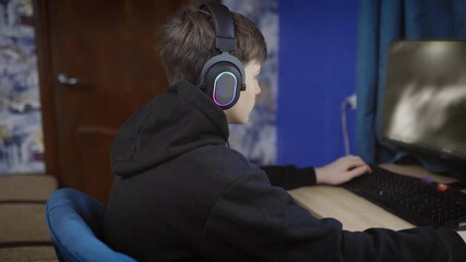Teenage boy in headset playing online first person shooter game on personal computer in room in apartment - Powered by Adobe