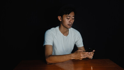 Young Asian Man Engaged with Smartphone in Dark Room. themes of digital addiction, late-night screen time, technology use, social media engagement, digital connection, isolation in the digital age.