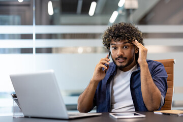 Worried young Indian man talking on the phone in the office, holding his head