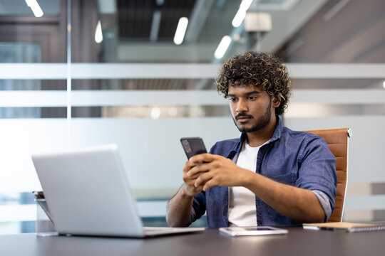 Serious young Indian man sitting in the lobby at his desk and using his mobile phone