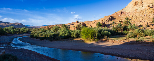 Scenic desert landscape with river, lush greenery, and distant mountains under a blue sky. Ait Benhaddou. Morocco