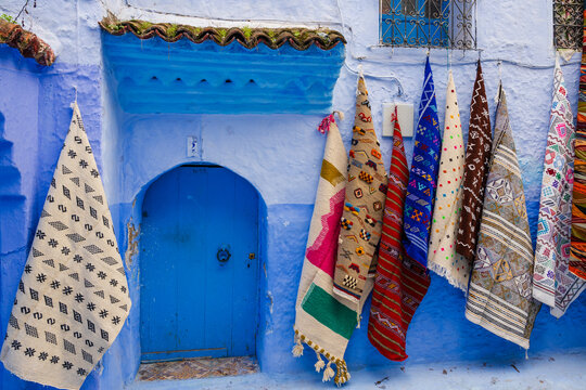 Colorful patterned rugs hang on a vibrant blue wall next to a turquoise door. Chefchaouen, Morocco