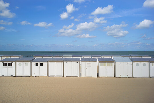White beach huts lined up on sandy shore under a blue sky with fluffy clouds. Blankenberge, West Flanders, Belgium