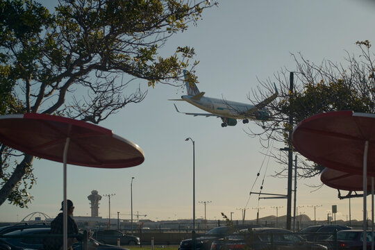Airplane landing over a parking lot with trees and red umbrella shades in the foreground. California, USA
