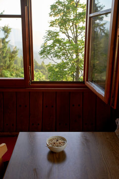 Fototapeta Bowl of cereal on wooden table by open window with scenic forest view. Salzkammergut, Dachstein Mountains, Austria