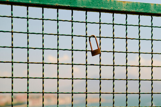 A rusted padlock hangs on a green wire fence with a blurred urban skyline in the background.