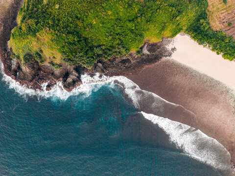 Aerial view of a lush green cliffside by the ocean and sandy beach with crashing waves. Lombok, Semeti, Indonesia