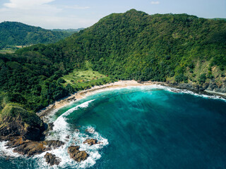 Aerial view of a secluded beach surrounded by lush green hills and turquoise ocean waters. Pantai Telawas Beach, Mekar Sari, Praya Barat, Central Lombok Regency, West Nusa Tenggara, Indonesia