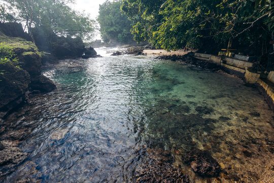 A tranquil, clear stream flows between lush green trees and rocks under an overcast sky. Ambon island, Maluku Islands, Indonesia