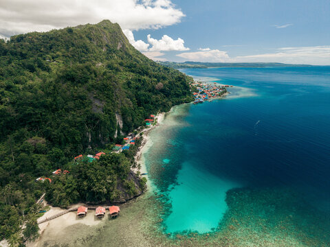 Aerial view of a tropical coastline with emerald waters and lush green hillside under a bright sky. Saleman Village, Seram Island, Indonesia