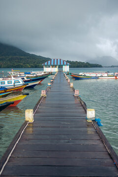 Wooden pier leading to boats on a cloudy day overlooking a serene mountain and lake. Banda Neira, Banda, Central Maluku, Indonesia