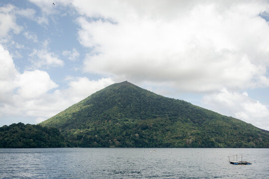 Lush green mountain under a cloudy sky with calm water in the foreground. Banda Neira, Banda, Central Maluku Regency, Indonesia