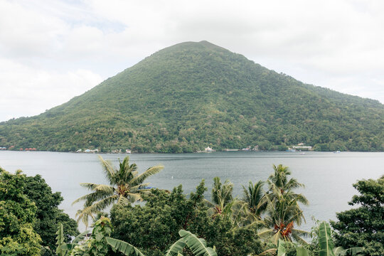 Lush green mountain rises above a calm bay with tropical trees in the foreground. Banda Neira, Banda, Central Maluku Regency, Indonesia