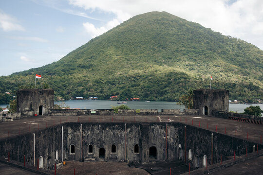 Ancient fort with weathered stone walls, lush green hill and calm water under a cloudy sky. Banda Neira, Banda, Central Maluku Regency, Indonesia