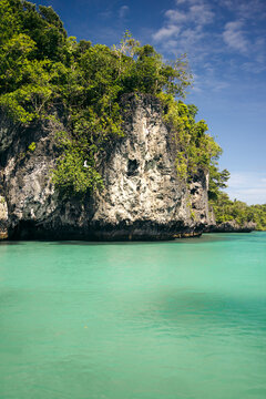 Turquoise waters surround a rocky cliff covered with lush green vegetation under a clear blue sky. Pulau Bair or Bair Island in Kei Kecil, Southeast Maluku Regency, Maluku, Indonesia