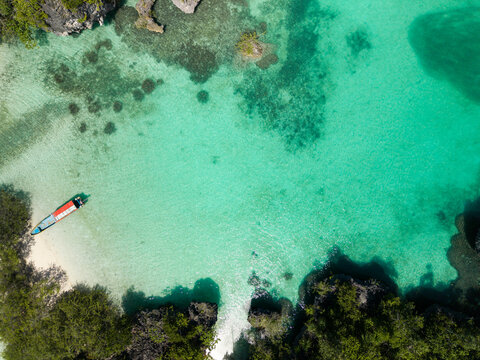 Aerial view of a turquoise blue lagoon with a small boat near a sandy shore surrounded by greenery. Pulau Bair or Bair Island in Kei Kecil, Southeast Maluku Regency, Indonesia