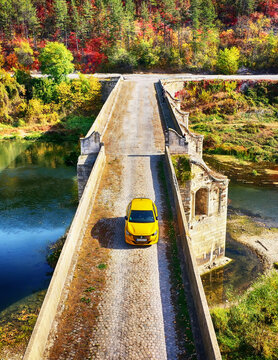 BYALA, BULGARIA - OCTOBER 28, 2025: Front view of a yellow Peugeot car on the Kolyo Ficheto Bridge over the Yantra River, framed by autumn forest and cobblestone road in warm sunlight.