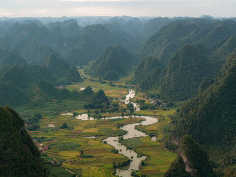 Aerial view of lush green valleys with winding river and distant hills under cloudy sky. Phong Nam, Tr˘ng Kh&middot;nh District, Cao Bang, Vietnam