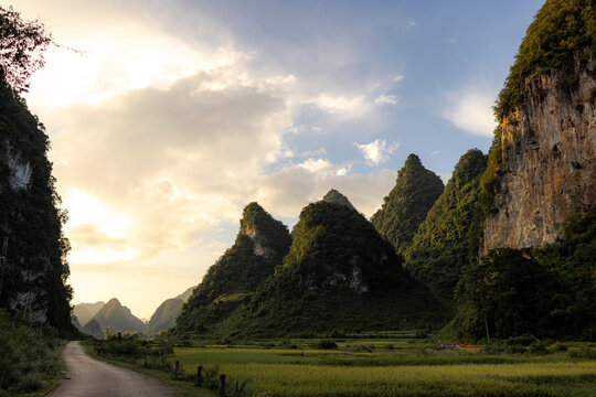 Dramatic mountains under a sky at sunset with lush green fields and a winding path. Phong Nam, Tr˘ng Kh·nh District, Cao Bang, Vietnam - Powered by Adobe