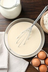 Liquid dough in bowl, whisk and ingredients on wooden table, flat lay