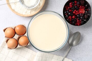 Liquid dough in bowl, berries and ingredients on light grey table, flat lay
