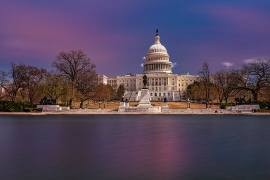 The US Capitol building at dusk with a vibrant sky and calm reflecting water in the foreground. Washington DC, WA, USA